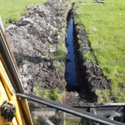 Wąski wykop wypełniony wodą, widok z kabiny koparki na łąkę z narzędziami w tle.