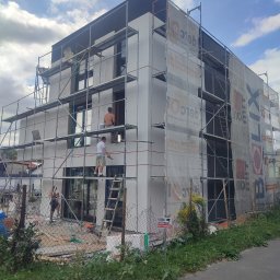 ŁUKASZ KOWALSKI Luk - Bud - Budynek w trakcie prac elewacyjnych, rusztowania, pracownicy w trakcie nakładania tynku. Część budynku osłonięta siatką ochronną z nadrukiem reklamowym.