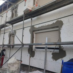 ŁUKASZ KOWALSKI Luk - Bud - Elewacja domu jednorodzinnego w trakcie prac, pokryta białymi płytami izolacyjnymi z czarnymi punktami. Metalowe rusztowanie, pracownik w czerwonych spodniach mocuje materiał. Wokół okien...