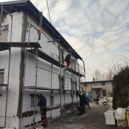 ŁUKASZ KOWALSKI Luk - Bud - Ekipa budowlana na rusztowaniu montuje białe płyty izolacyjne na ścianach budynku, częściowo pokryte szarą zaprawą. Widoczne okna z roletami, pochmurne niebo.