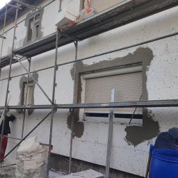 ŁUKASZ KOWALSKI Luk - Bud - Elewacja budynku w trakcie ocieplania białym styropianem, z widocznym rusztowaniem i dwoma pracownikami nakładającymi szary tynk wokół okien z roletami.