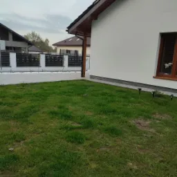 Boczny widok białego domu z brązowymi ramami okiennymi i dachem, widoczny trawnik i fragment ogrodzenia z białymi słupkami i czarnymi przęsłami.