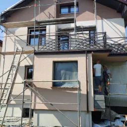 Dwóch mężczyzn na rusztowaniu przy dwukondygnacyjnym domu, jeden maluje fragment elewacji w kolorze beżowym, okno zabezpieczone folią, widoczna drabina i fragment chodnika z kostki.