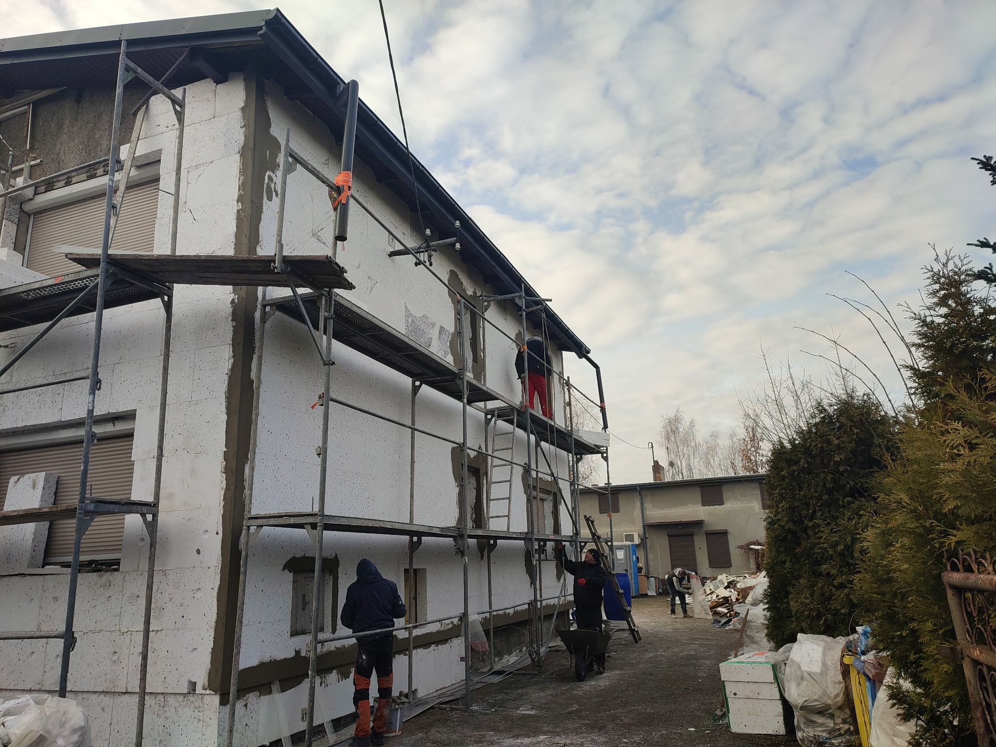 Dom w trakcie ocieplania styropianem, widoczne rusztowania i pracownicy. Częściowo nałożony klej na styropian. Budynek z szarego pustaka.