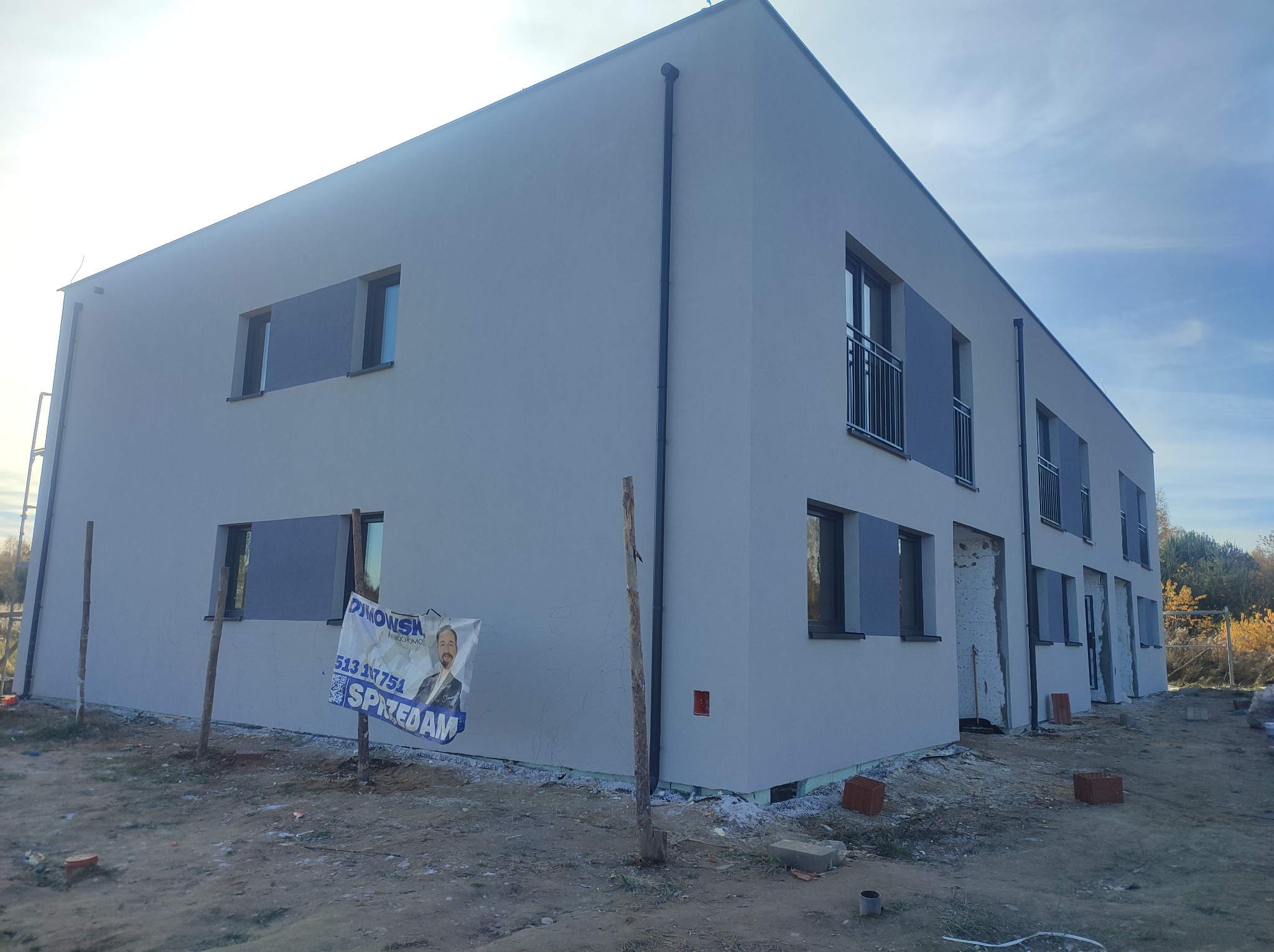 Dwukondygnacyjny dom w trakcie wykańczania elewacji, z widocznym banerem reklamowym nieruchomości na pierwszym planie i fragmentami ocieplenia przy gruncie.
