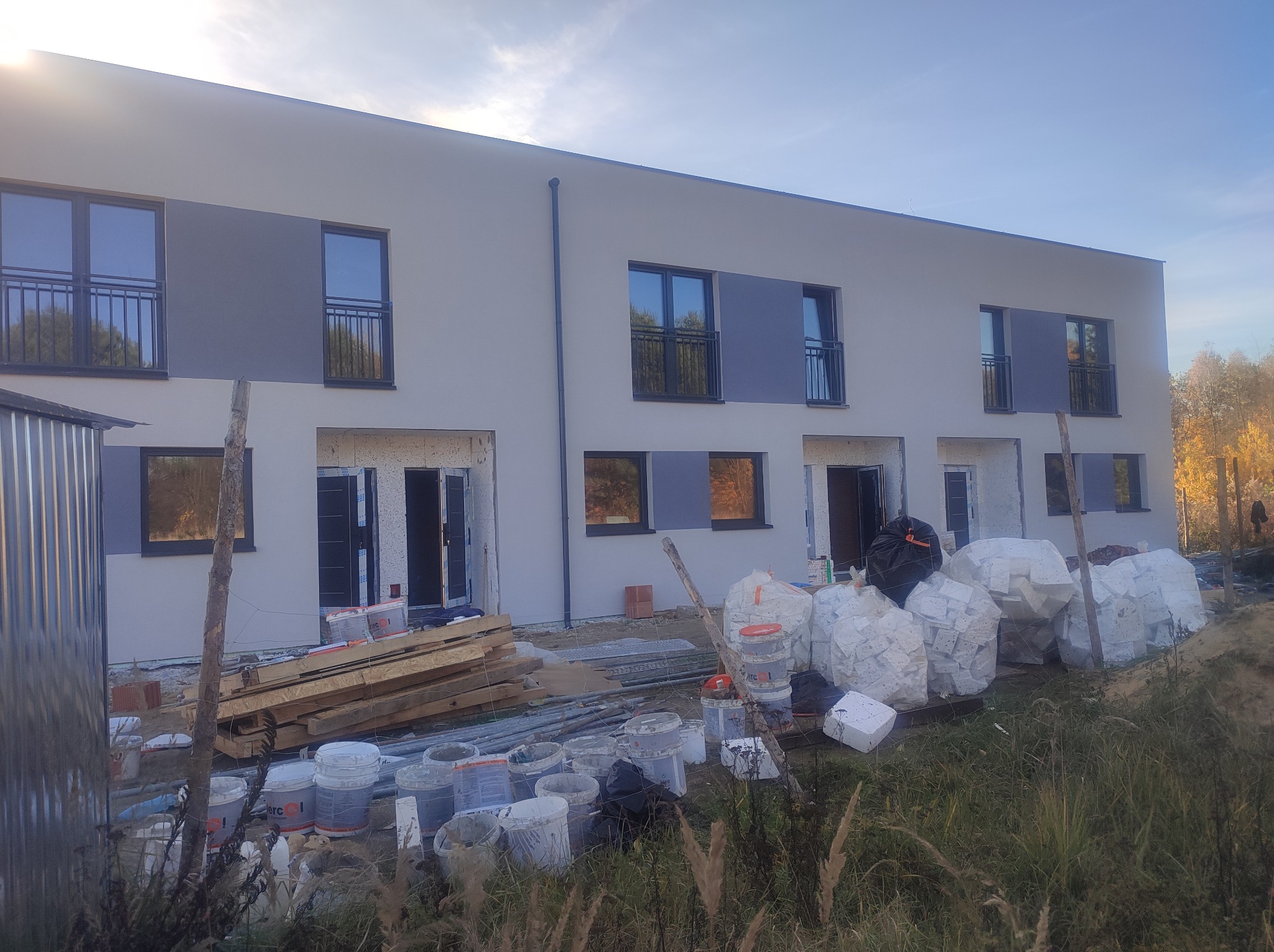 Dwukondygnacyjny budynek w trakcie prac elewacyjnych, widoczne materiały budowlane i rusztowania. Częściowo otynkowana fasada z oknami i balkonami.