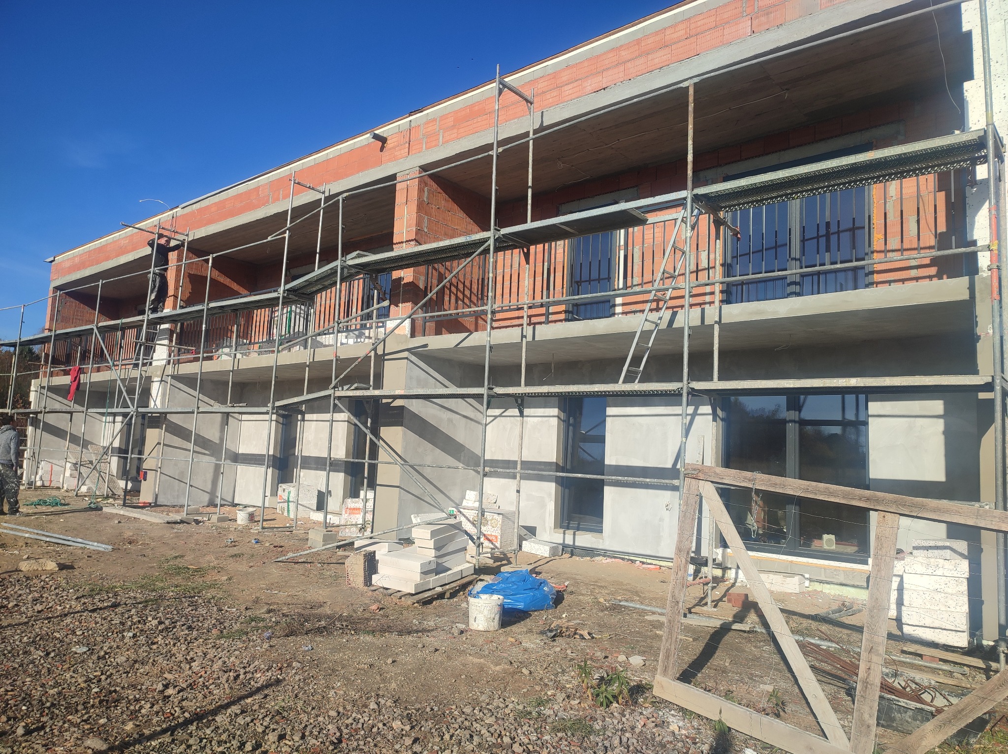 Budynek w trakcie tynkowania elewacji, widoczne rusztowania, częściowo otynkowane ściany, okna i materiały budowlane na placu budowy w słoneczny dzień.