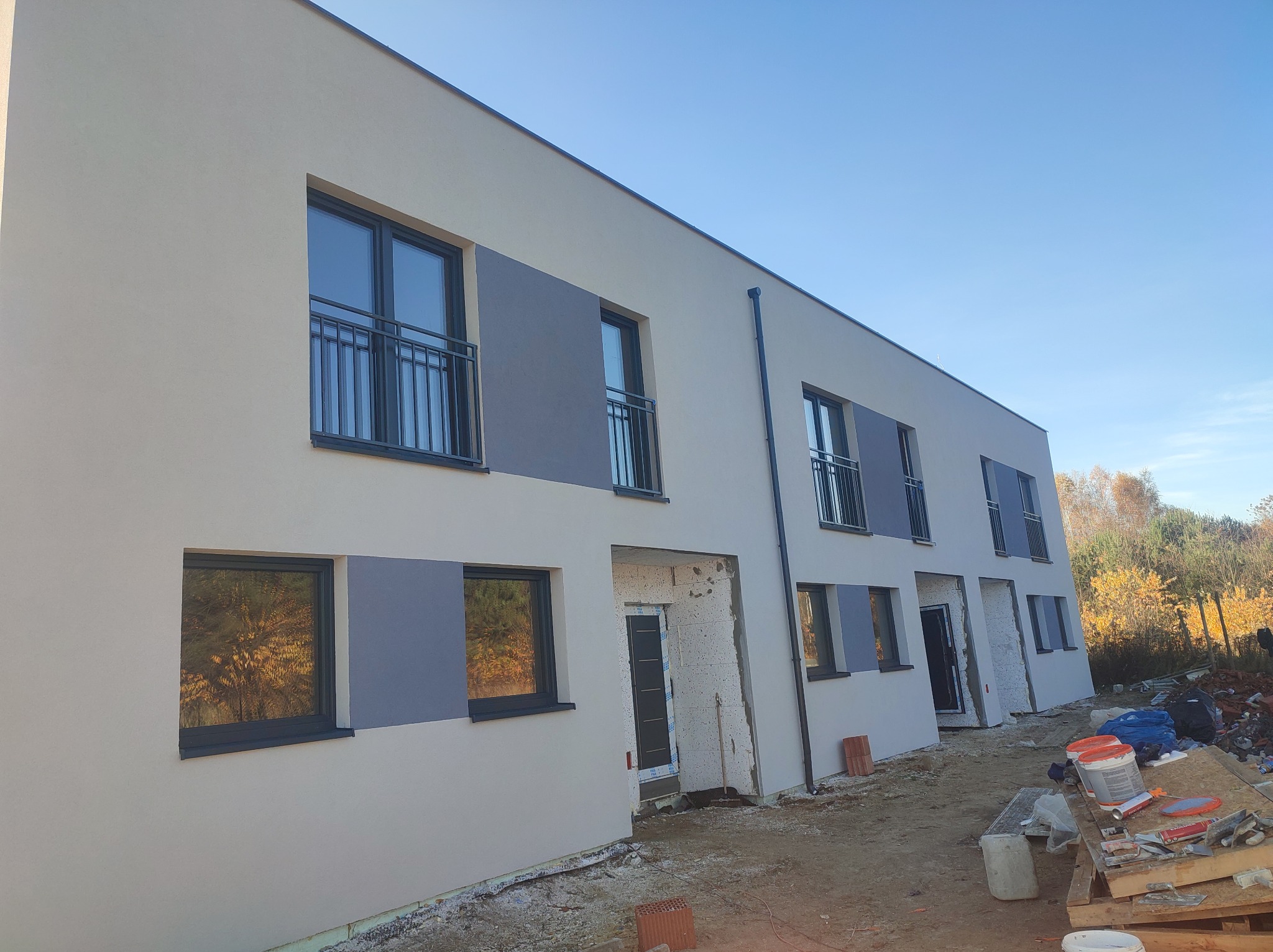 Nowoczesny budynek wielorodzinny z jasną elewacją i szarymi akcentami. Widoczne okna z balkonami i częściowo wykończone wejścia. Prace wykończeniowe w toku.