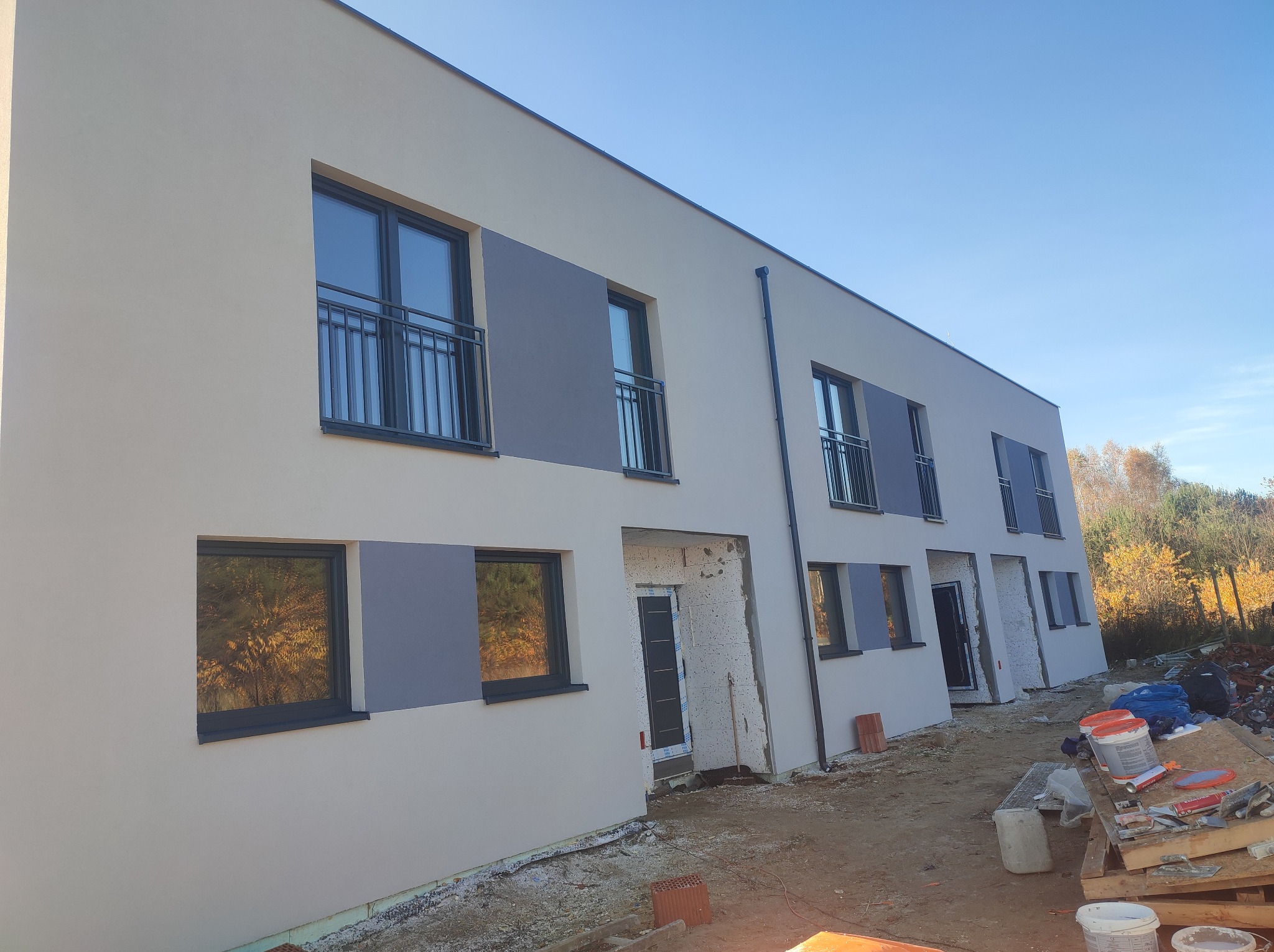 Nowoczesny budynek w trakcie prac elewacyjnych: jasna fasada z szarymi akcentami, czarne okna z balkonami, widoczne materiały budowlane i teren budowy.