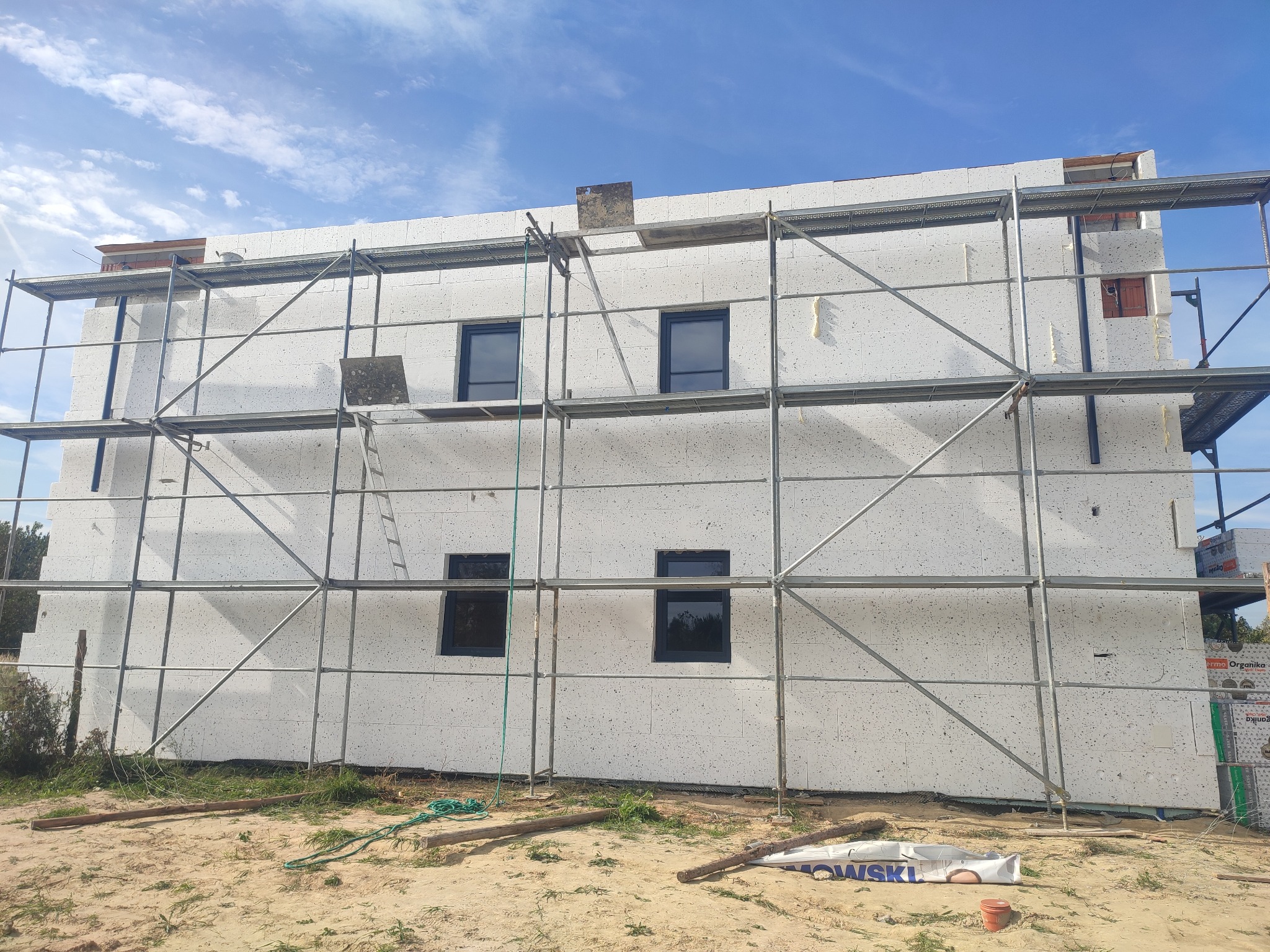 Budynek w trakcie ocieplania, widoczne rusztowania i białe płyty izolacyjne. Okna zamontowane, nieotynkowana elewacja z widocznymi szczelinami między płytami.