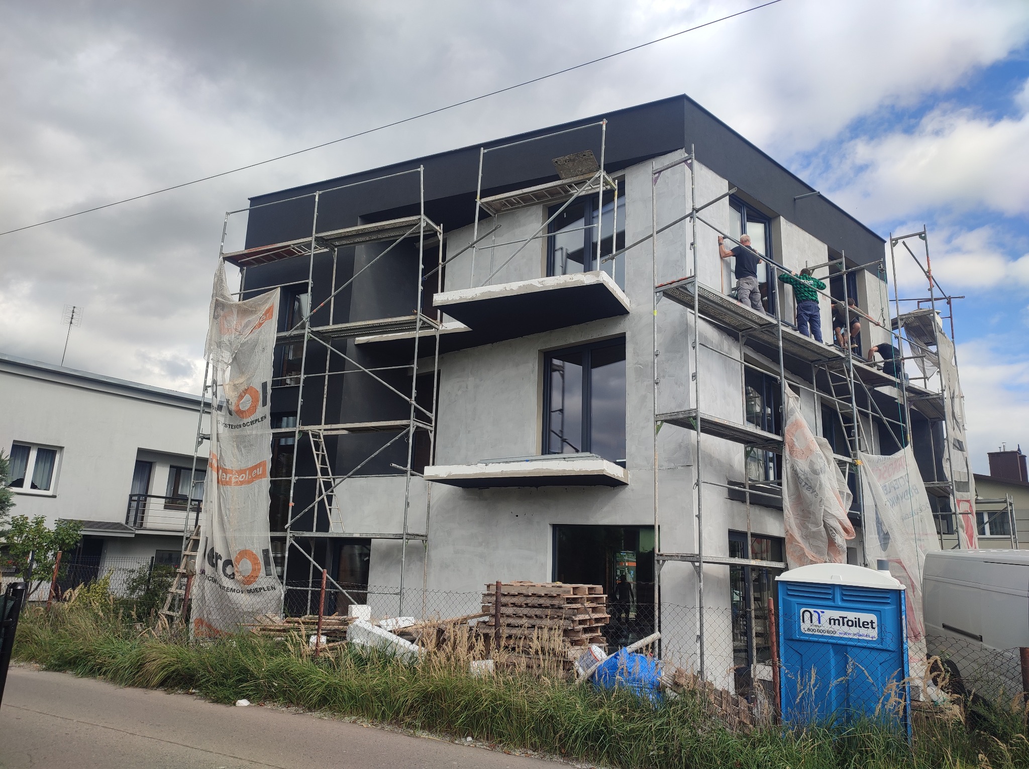 Wielopiętrowy budynek w trakcie prac elewacyjnych. Widoczne rusztowania, pracownicy oraz materiały budowlane. Na pierwszym planie toaleta przenośna.