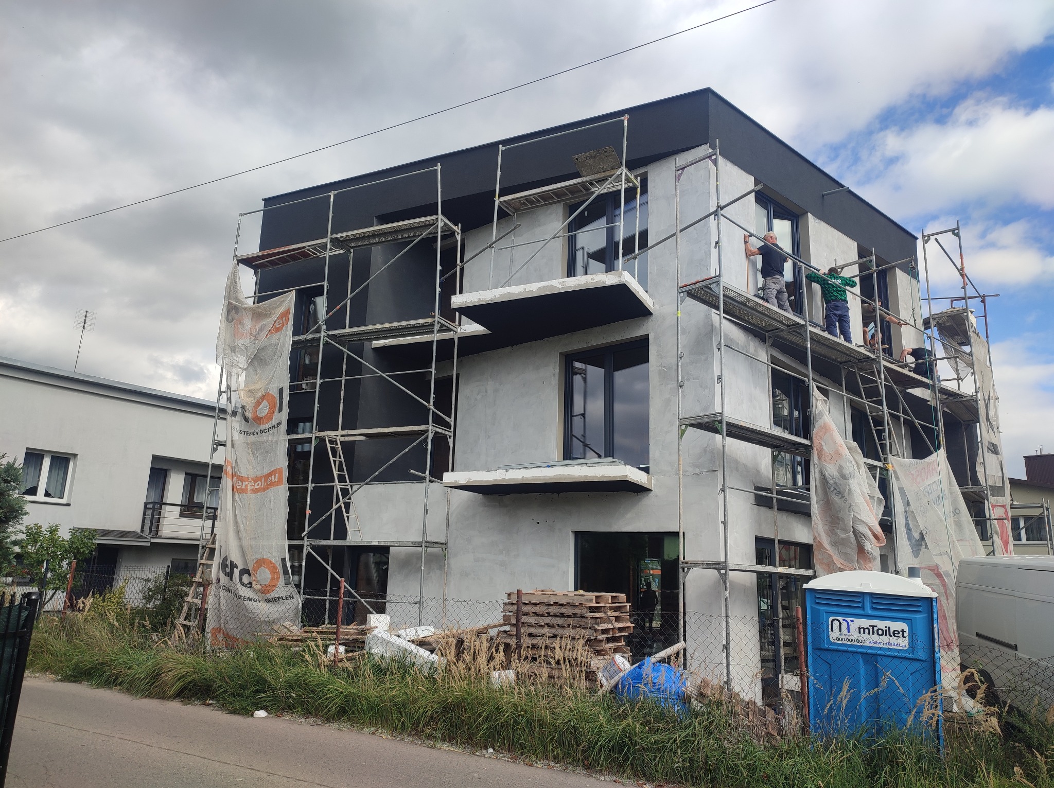 Budynek w trakcie tynkowania elewacji, widoczne rusztowania, pracownicy i przenośna toaleta. Szare niebo w tle. Częściowo otynkowana ściana budynku.