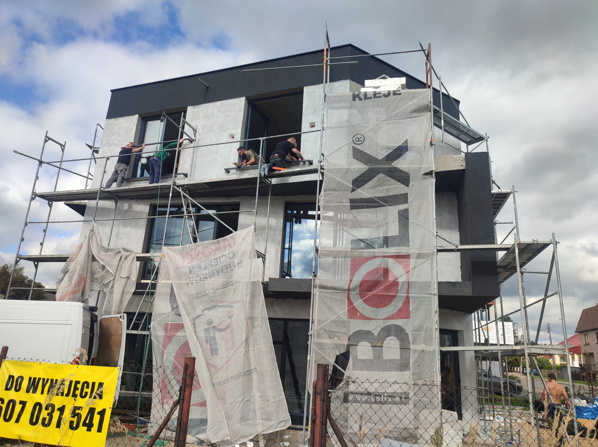 Dom w trakcie prac elewacyjnych: robotnicy na rusztowaniu montują okna, elewacja częściowo pokryta tynkiem, zabezpieczona siatką z logo producenta. Widoczny baner 'Do wynajęcia'.