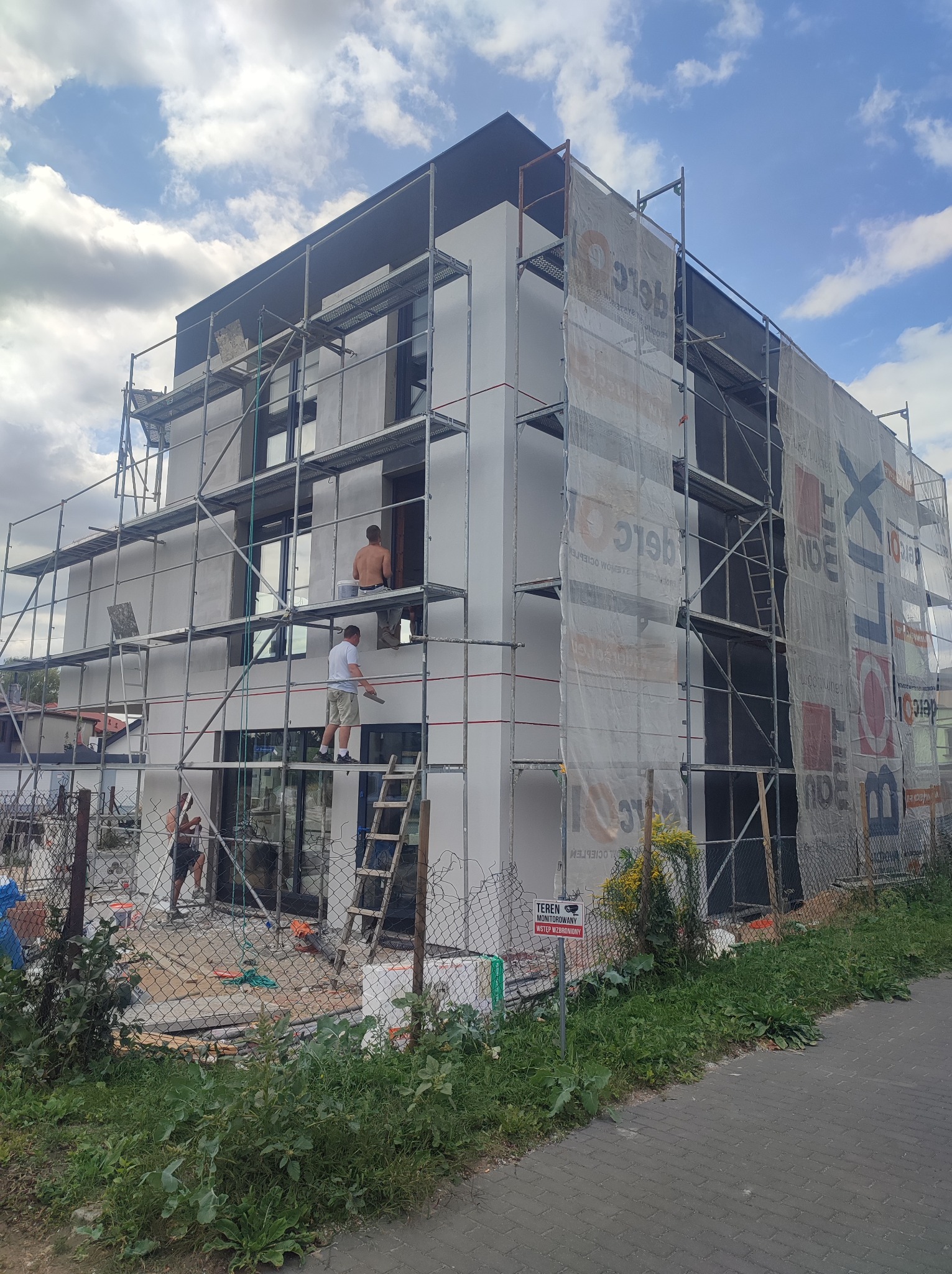 Budynek w trakcie prac elewacyjnych, rusztowania, pracownicy w trakcie nakładania tynku. Część budynku osłonięta siatką ochronną z nadrukiem reklamowym.