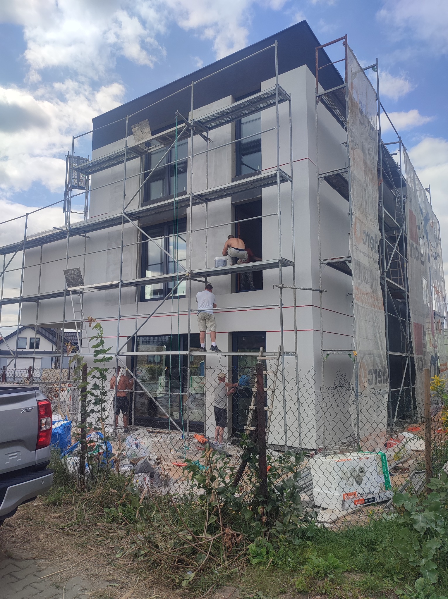 Ocieplanie elewacji domu jednorodzinnego z użyciem rusztowań. Widoczni pracownicy podczas wykonywania prac. Częściowo zakryte rusztowania siatką ochronną.