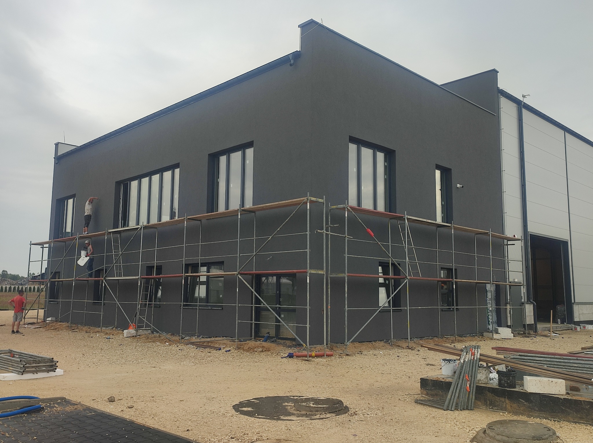 Szara elewacja budynku w trakcie prac wykończeniowych. Widoczne rusztowania, pracownicy i materiały budowlane. Część budynku w kolorze białym.