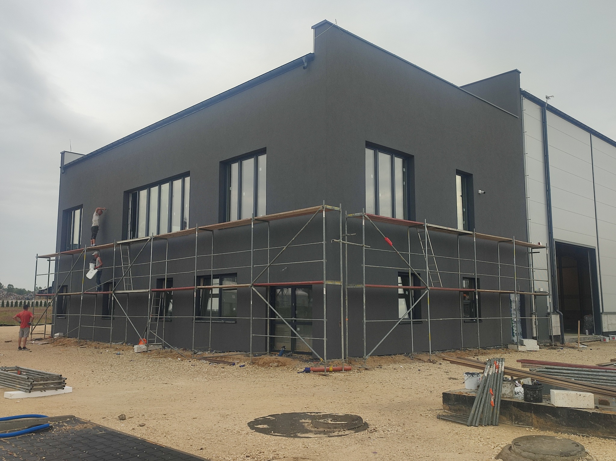 Budynek w trakcie tynkowania na szaro, z rusztowaniami i pracownikiem na wysokości. Widoczne okna i elementy konstrukcyjne. Praca w toku, teren budowy.