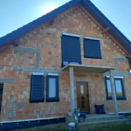 Niedokończony dom jednorodzinny z cegły z zamontowanymi roletami zewnętrznymi i oknami z widocznym ociepleniem fundamentów, czekający na elewację.