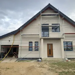 Nowoczesny dom jednorodzinny w trakcie prac elewacyjnych, widoczne rusztowania, świeżo otynkowana elewacja w jasnym kolorze, czarna dachówka, teren wokół domu częściowo zagospodarowany.