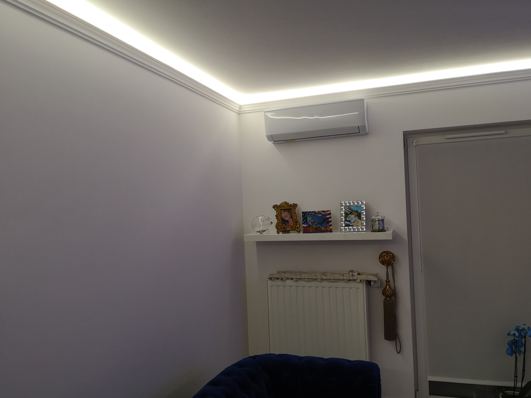 Nowoczesny, biały klimatyzator ścienny w jasnym salonie z oświetleniem LED podsufitowym, półką z dekoracjami i niebieskim fotelem w tle.