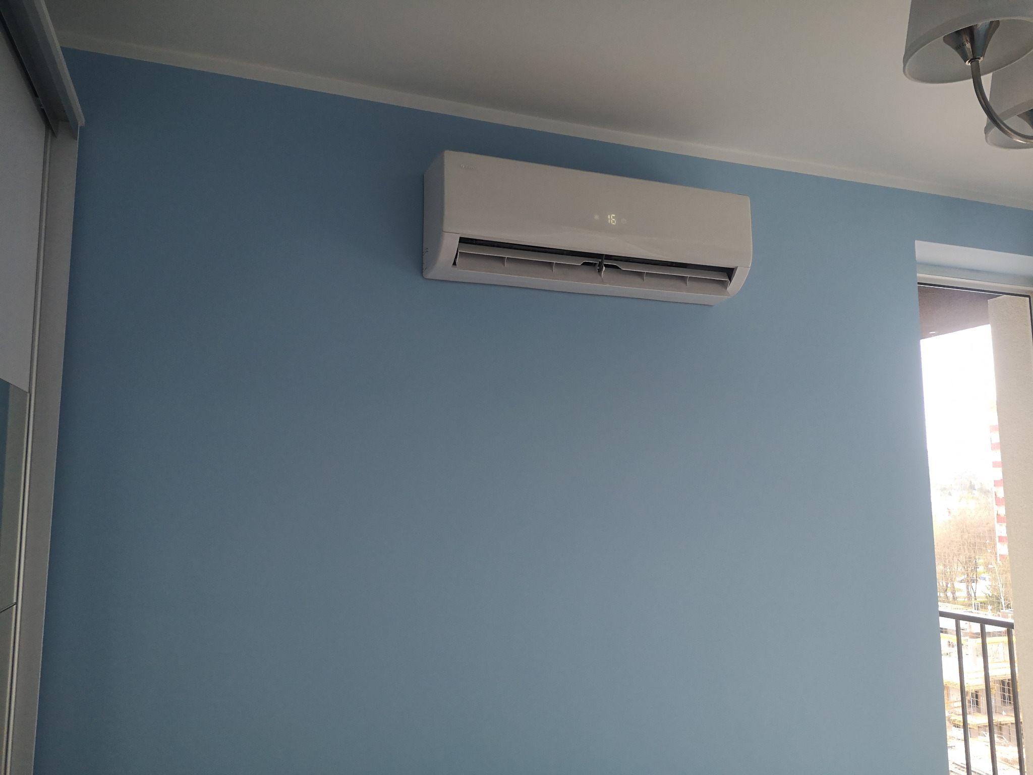 Biały klimatyzator Gree Multi Split zamontowany na błękitnej ścianie w pokoju, widoczny wyświetlacz z temperaturą 16 stopni Celsjusza, w tle okno balkonowe.