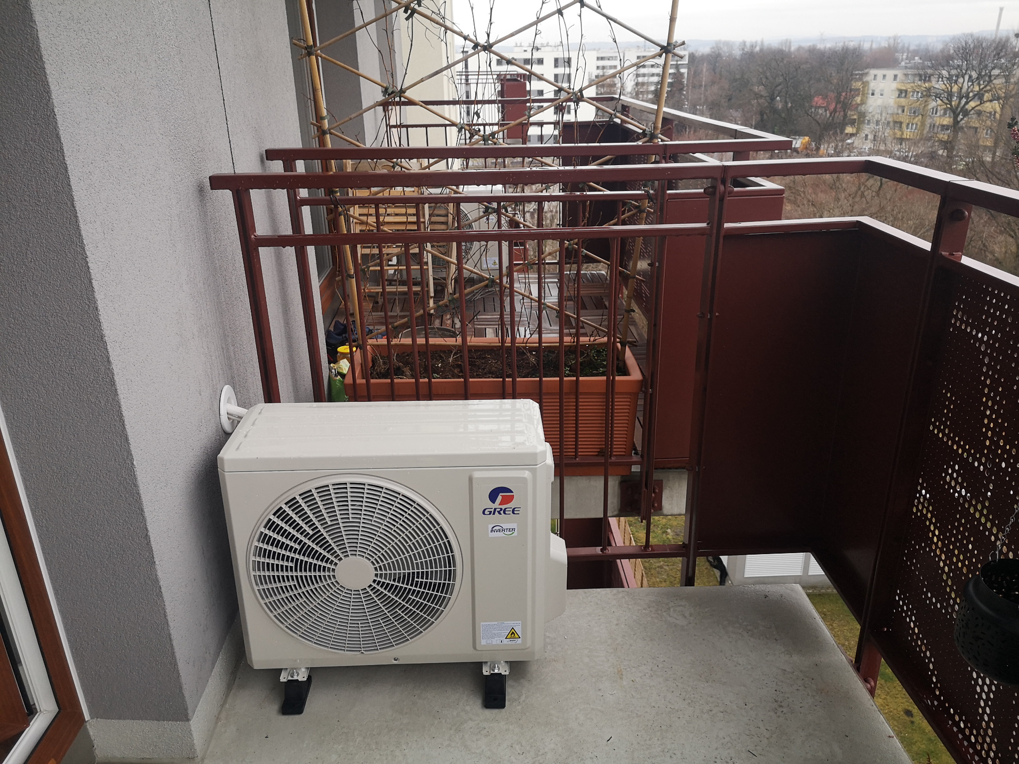 Jednostka zewnętrzna klimatyzatora marki Gree zamontowana na balkonie, widok z boku na szarym tle ściany i brązowej balustrady.