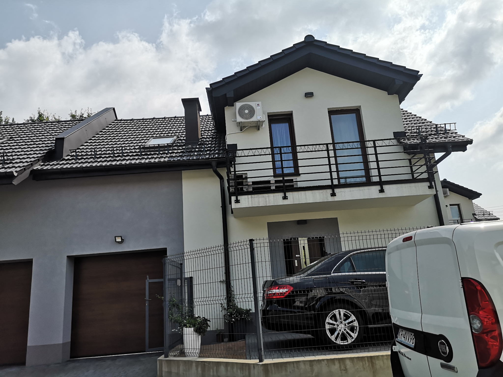 Biały dom z czarnym dachem, balkonem i zamontowaną jednostką klimatyzacji na ścianie, widoczny samochód osobowy i dostawczy na podjeździe.