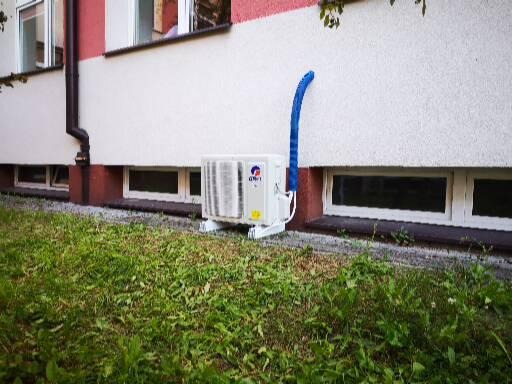 Zewnętrzna jednostka klimatyzacji zamontowana przy ścianie budynku z widocznym niebieskim przewodem odprowadzającym skropliny, trawnik w pierwszym planie.