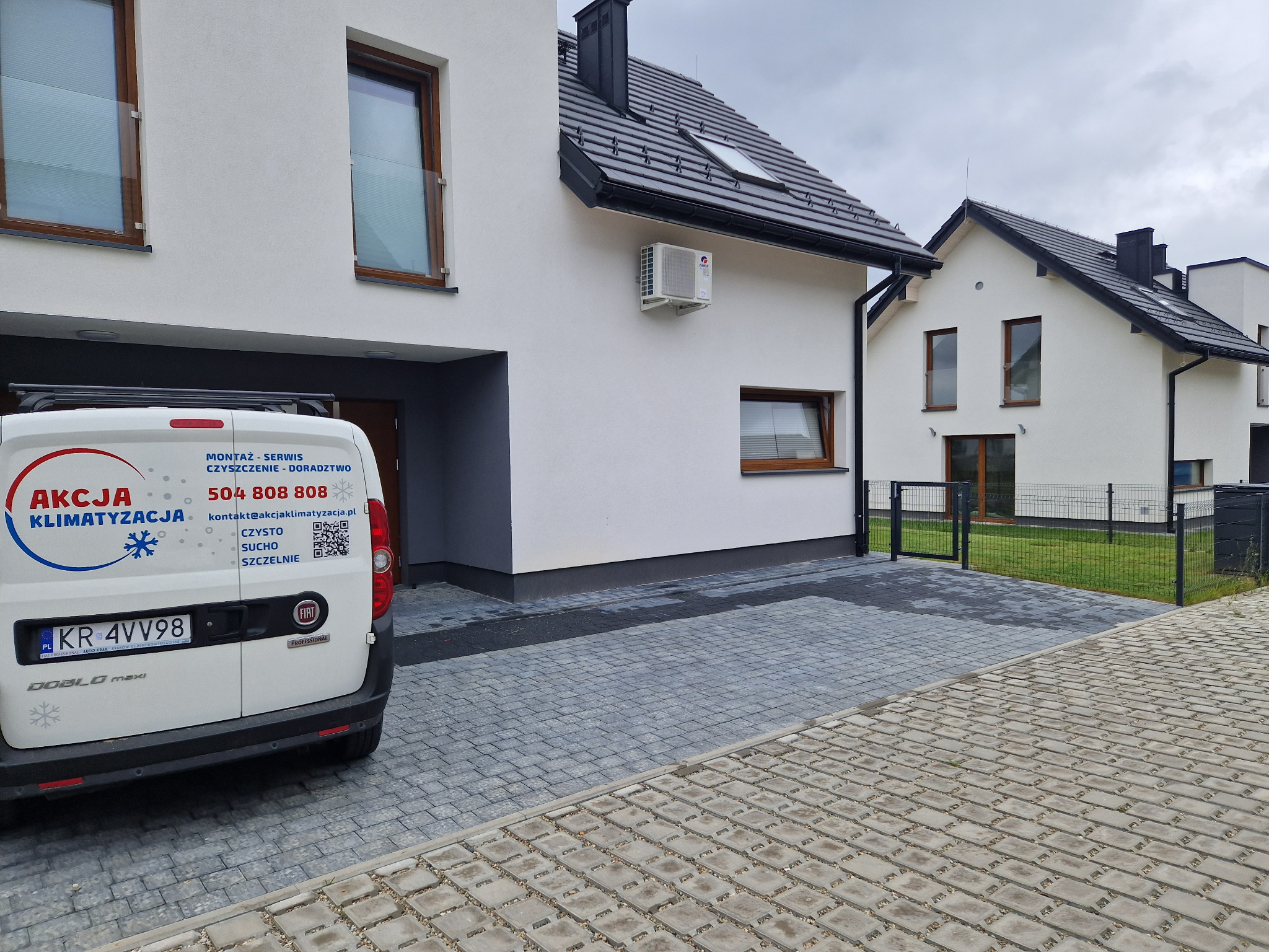 Biały budynek jednorodzinny z zamontowaną klimatyzacją zewnętrzną, widoczny z boku samochód dostawczy z reklamą firmy oferującej montaż i serwis klimatyzacji.