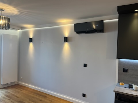 Czarny klimatyzator ścienny zamontowany w nowoczesnym salonie z drewnianą podłogą, oświetleniem LED i czarnymi kinkietami.