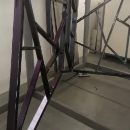 Metalowa konstrukcja pomalowana na ciemny fiolet, zawieszona na haku w lakierni proszkowej, widoczny fragment linii technologicznej i tablica BHP w tle.