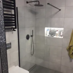 Nowoczesna łazienka z szarymi płytkami, kabiną prysznicową typu walk-in z czarnymi elementami, grzejnikiem drabinkowym, podświetlaną wnęką z kosmetykami i wiszącą toaletą.