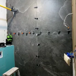Montaż wielkoformatowych, grafitowych płytek imitujących kamień na ścianie. Użyto poziomicy laserowej i klipsów poziomujących. Widoczna drabina malarska i niezakończone prace elektryczne.