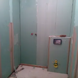 Łazienka w trakcie remontu w Dębicy: ściany wyłożone płytami kartonowo-gipsowymi, widoczne otwory na instalacje, fragment podłogi i szczotka na górnej krawędzi zabudowy stelaża podtynkowego.