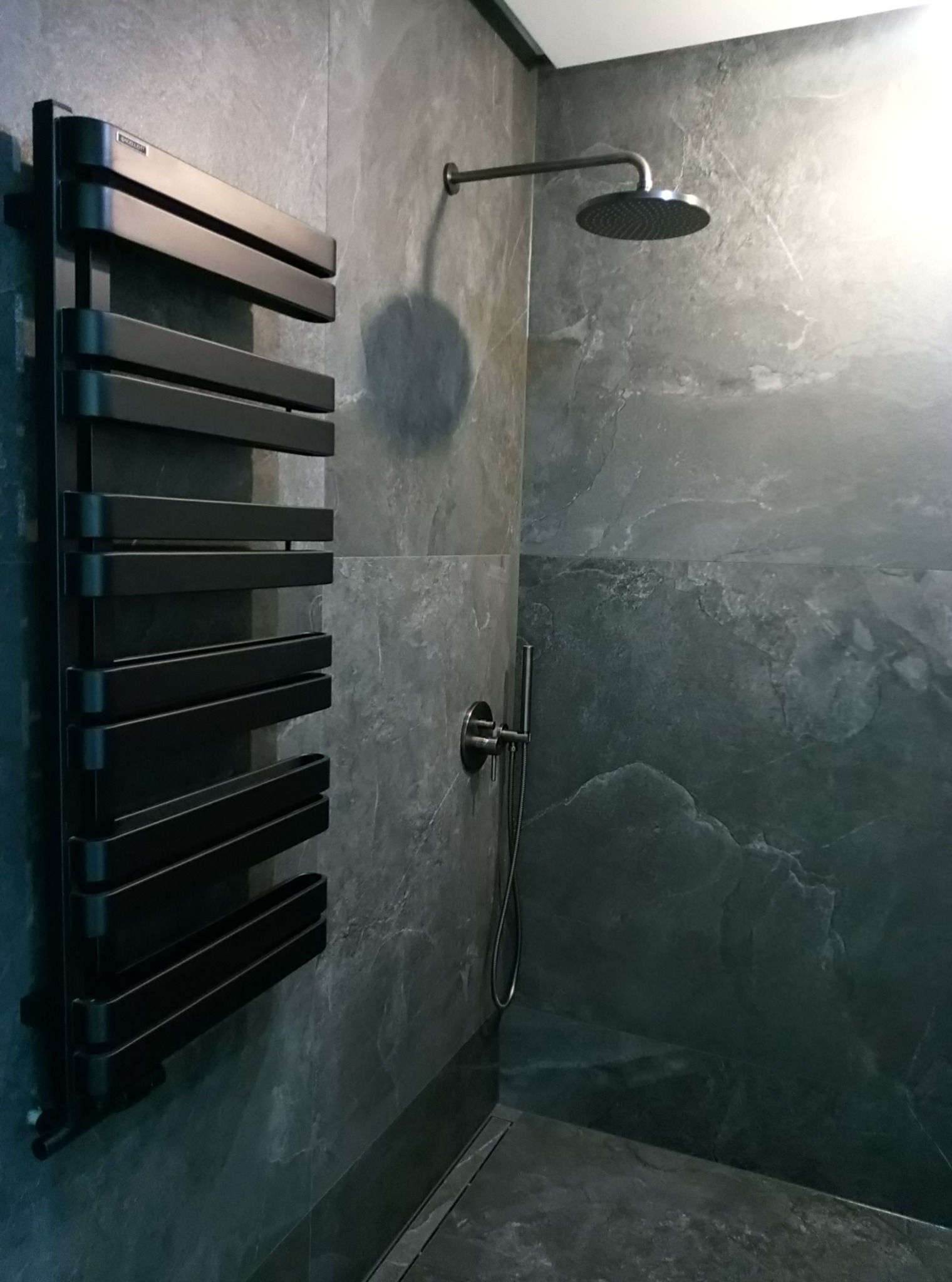 Nowoczesna kabina prysznicowa z deszczownicą i czarnym grzejnikiem drabinkowym na tle szarych, marmurkowych płytek.
