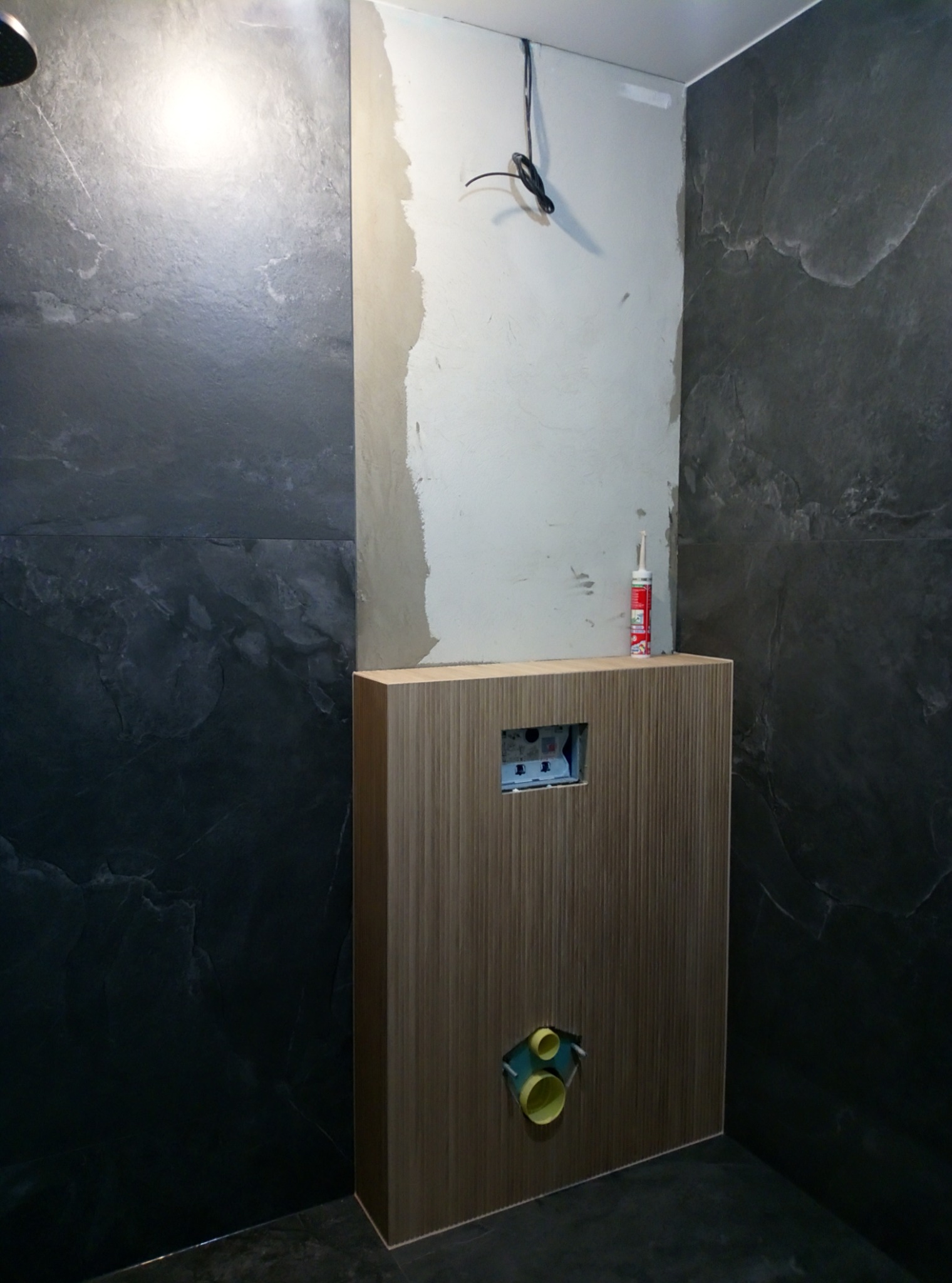 Częściowo wykończona łazienka z ciemnymi płytkami imitującymi kamień, widoczna zabudowa stelaża WC z otworami na rury odpływowe, ściana przygotowana pod płytki z widocznymi kablami elektrycznymi...