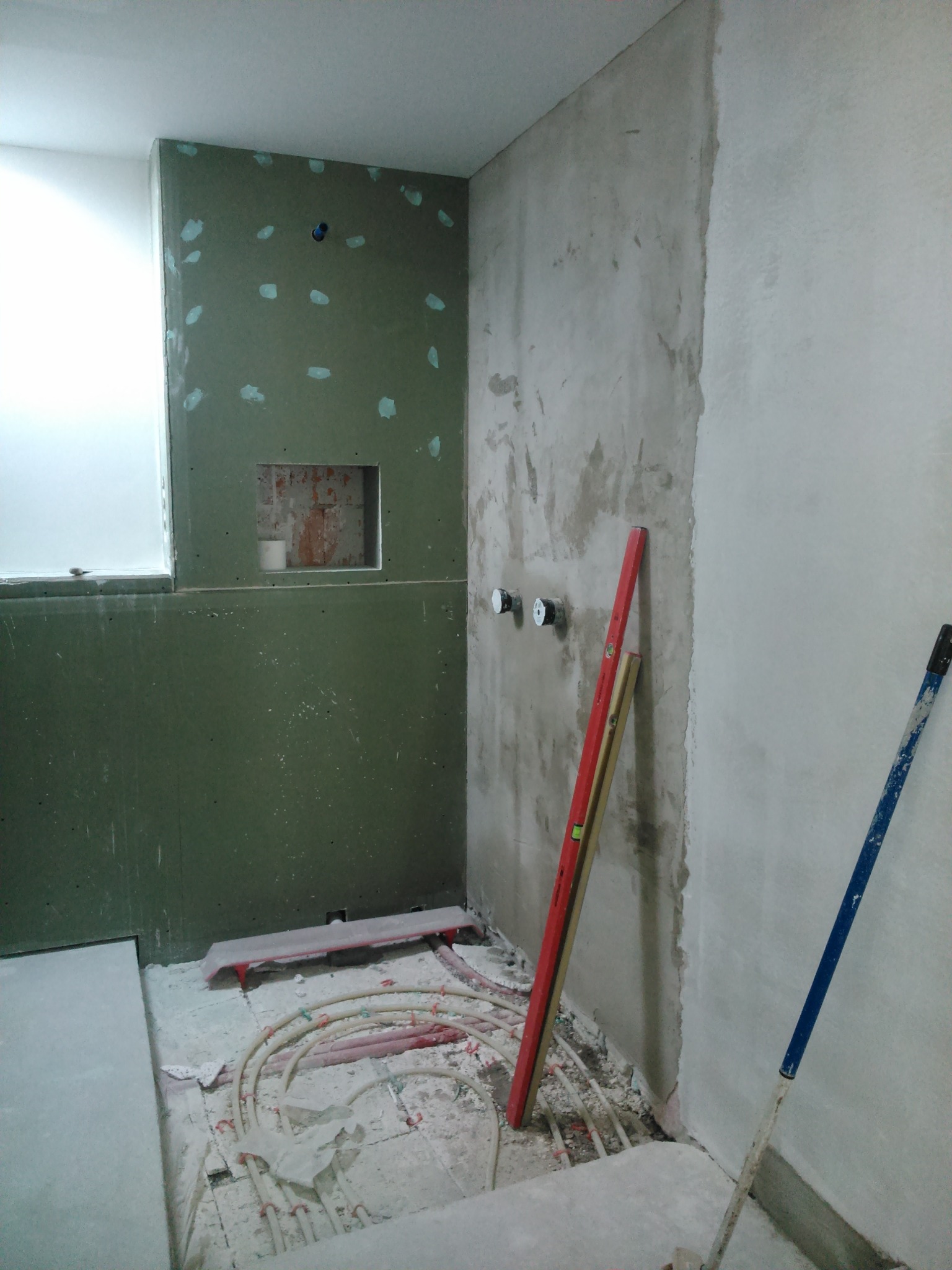 Łazienka w trakcie remontu: ściana z płyt karton gips, rury ogrzewania podłogowego, poziomice oparta o ścianę, widoczne przyłącza wodne.