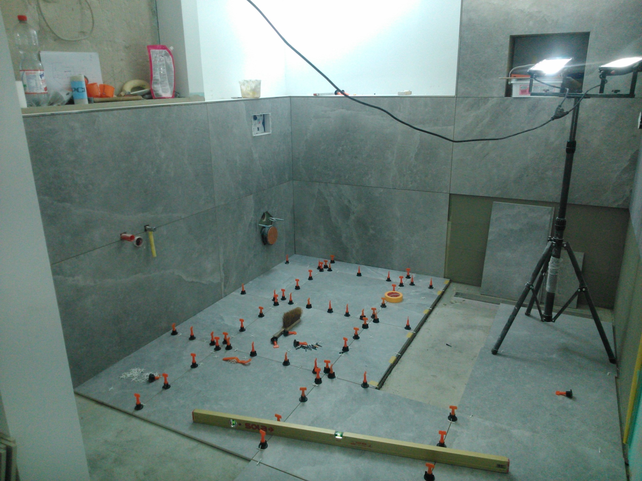 Układanie szarych płytek podłogowych w łazience, widoczne poziomica Sola, system poziomowania płytek z pomarańczowymi klipsami, szczotka do fugowania i lampy na statywie.