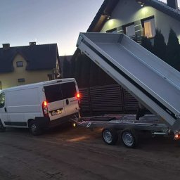 Łukasz Federowicz - Biały bus z podpiętą przyczepą wywrotką na tle domów jednorodzinnych o zmierzchu. Przyczepa w pozycji podniesionej, gotowa do wyładunku.