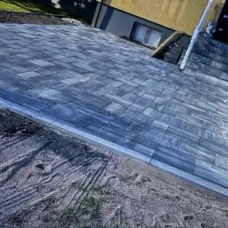 Łukasz Federowicz - Ułożona, szara kostka brukowa przed domem z oświetleniem, widoczne ślady opon na ziemi. Kąt ujęcia z dołu podkreśla perspektywę i wzór kostki.