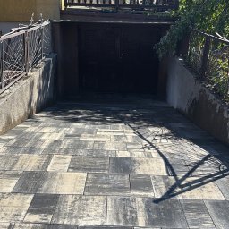 Łukasz Federowicz - Ułożona kostka brukowa w odcieniach szarości i beżu prowadząca do garażu w budynku z balkonem. Cień metalowego ogrodzenia na kostce. Słoneczny dzień.