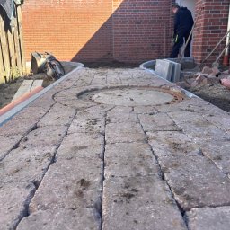 Łukasz Federowicz - Układanie kostki brukowej z okrągłym elementem centralnym. Widoczne narzędzia i materiały budowlane. Prace w toku, częściowo ułożona kostka, ceglana ściana w tle.