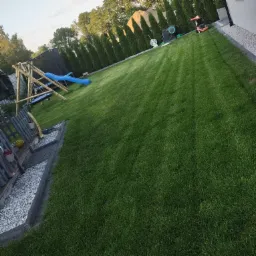 Perfekcyjnie przystrzyżony trawnik z równymi pasami, drewniany plac zabaw z niebieską zjeżdżalnią, rząd tuj w tle, obrzeża z kostki brukowej i białego grysu.