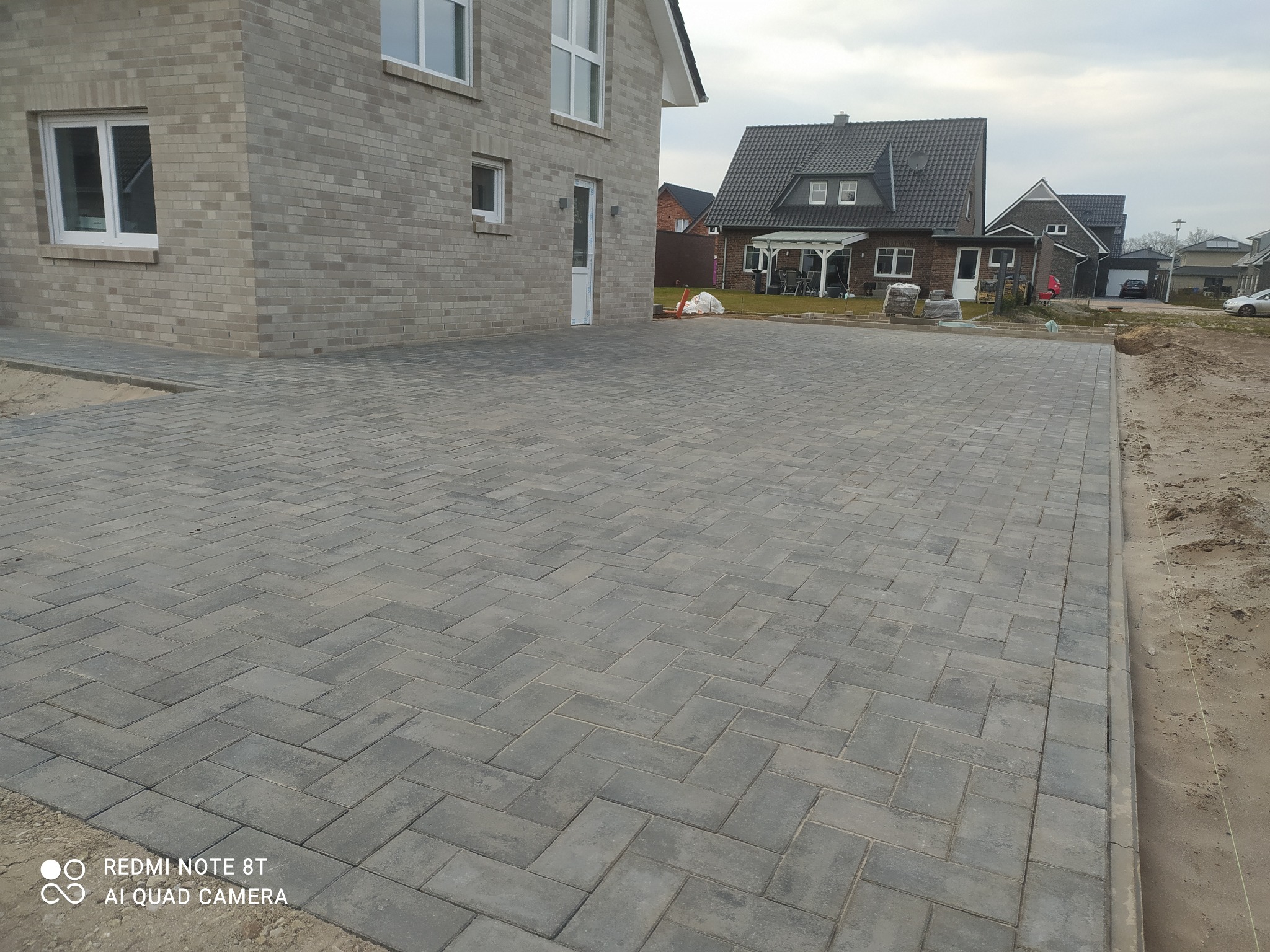 Ułożona szara kostka brukowa w jodełkę przed nowoczesnym domem z szarej cegły, widoczny piasek po bokach, w tle inne domy.