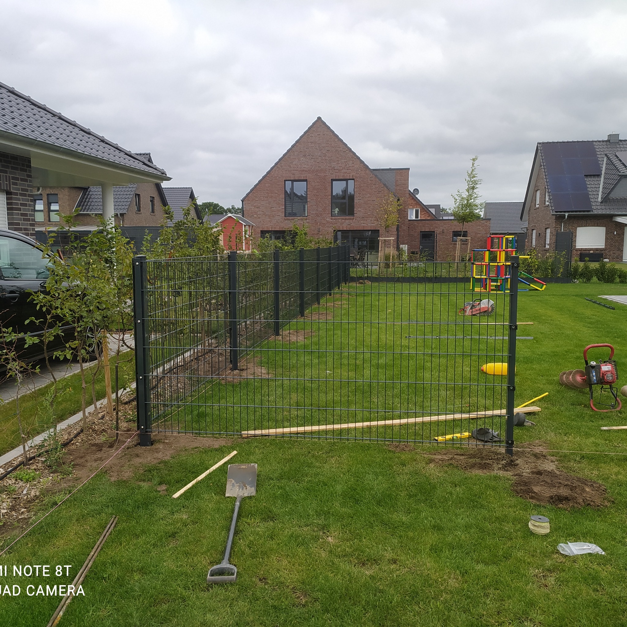 Montaż ogrodzenia panelowego na trawniku, widoczne narzędzia: łopata, poziomica, sznurek, w tle domy jednorodzinne i plac zabaw.