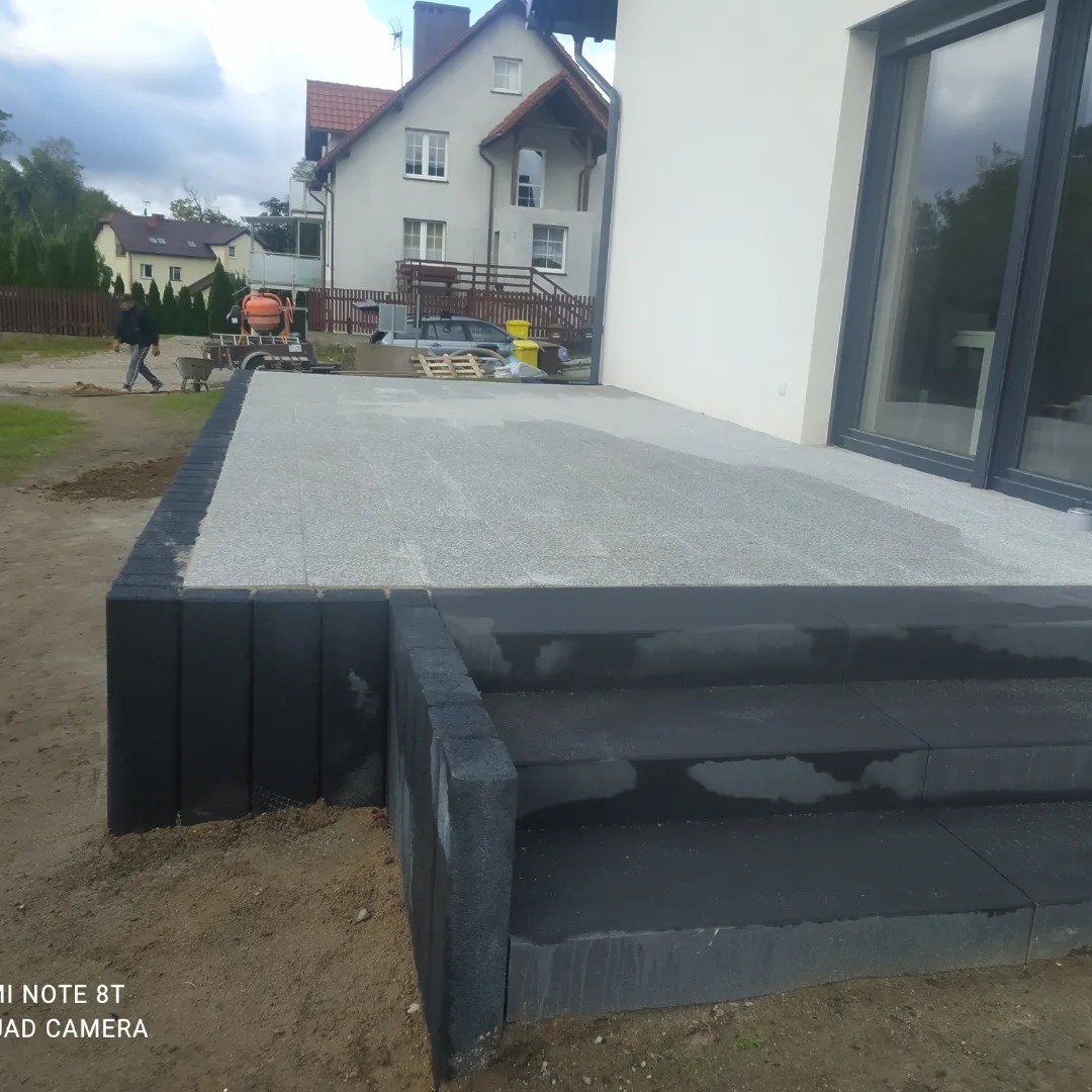 Nowo ułożony taras z jasnoszarej kostki brukowej z czarnymi obrzeżami i schodami, widoczny betoniarka i pracownik w tle.