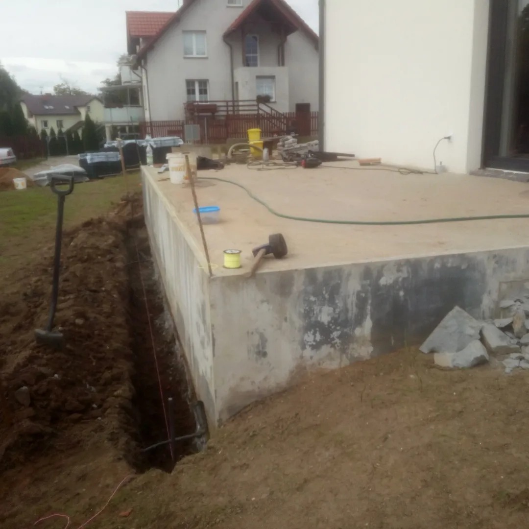 Wykop pod fundament z widoczną linią wyznaczoną sznurkiem, obok betonowa wylewka fundamentu i narzędzia budowlane, w tle dom jednorodzinny.