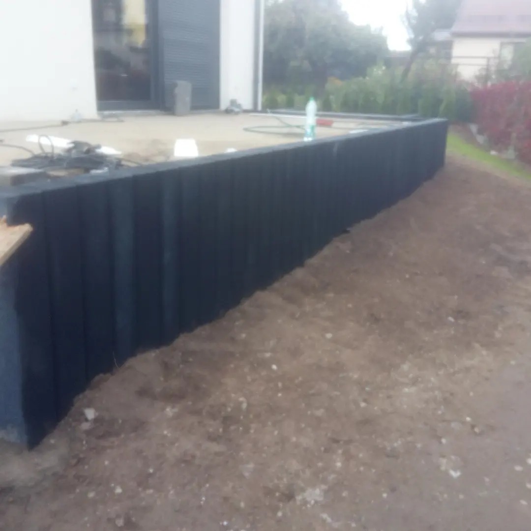 Ciemnoszara, pionowo żebrowana obudowa wykonana z tworzywa sztucznego, prawdopodobnie szamba, w trakcie montażu, widoczna na tle domu z szarymi roletami i przygotowanego terenu.