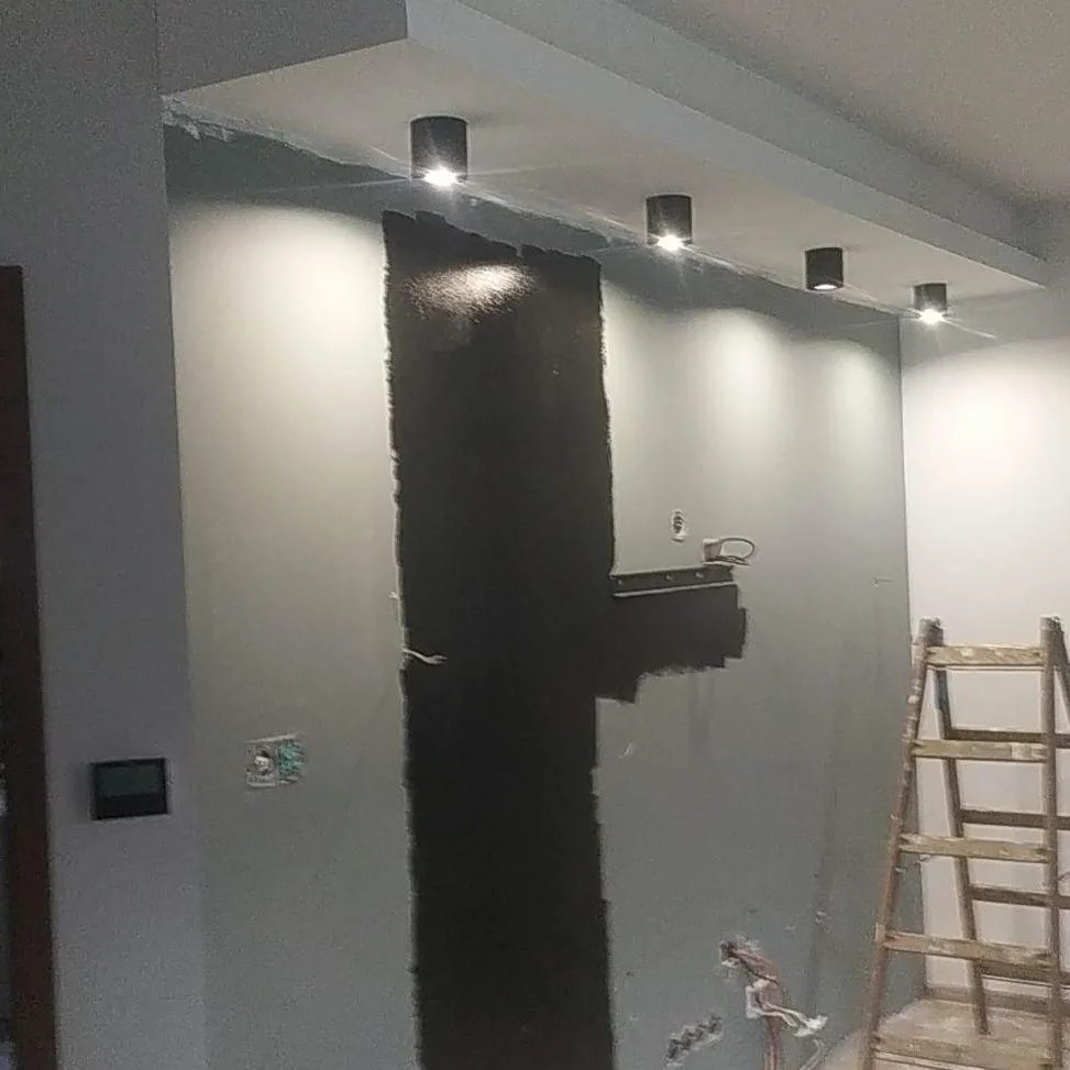 Ściana w trakcie malowania na szaro, z pionowym pasem świeżo nałożonej czarnej farby, widoczne puszki elektryczne i drabina drewniana, oświetlenie sufitowe punktowe.
