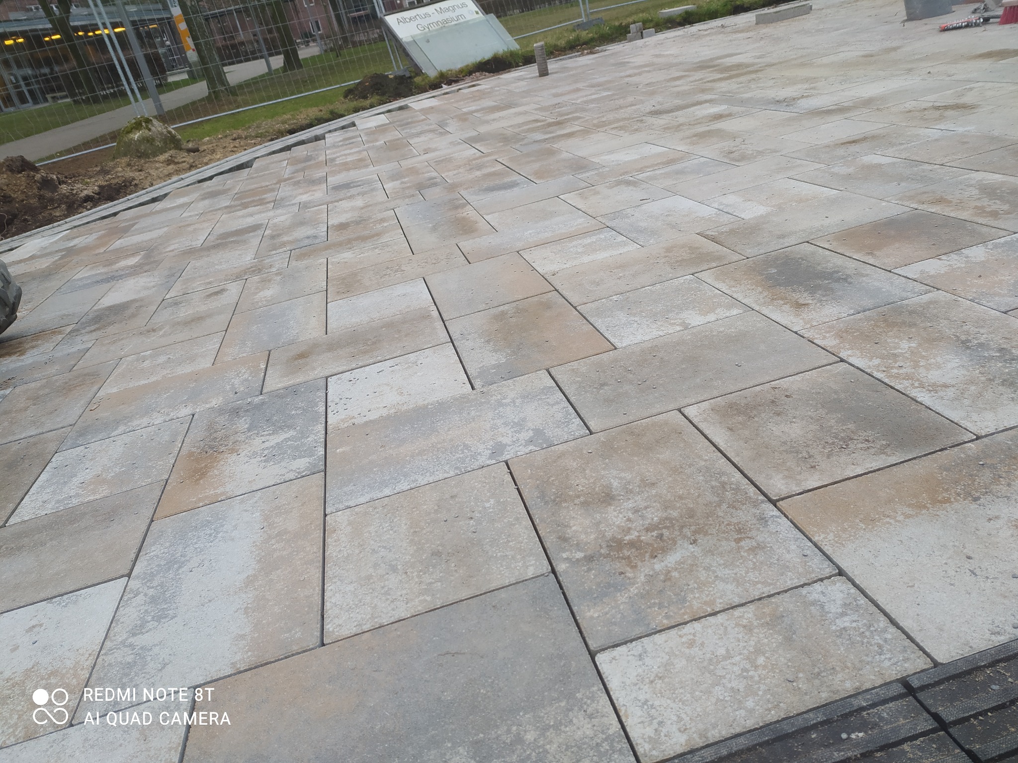 Ułożona kostka brukowa w odcieniach szarości i beżu, widok z góry na plac w trakcie prac wykończeniowych, w tle budynek z ogrodzeniem.